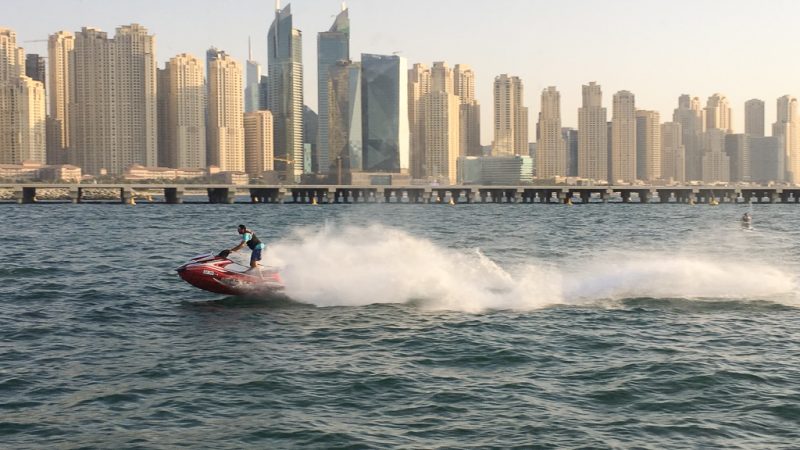 Photo taken across water to the skyscrapers of the Dubai skyline. A man is powering a jet ski at speed across the middle of the picture