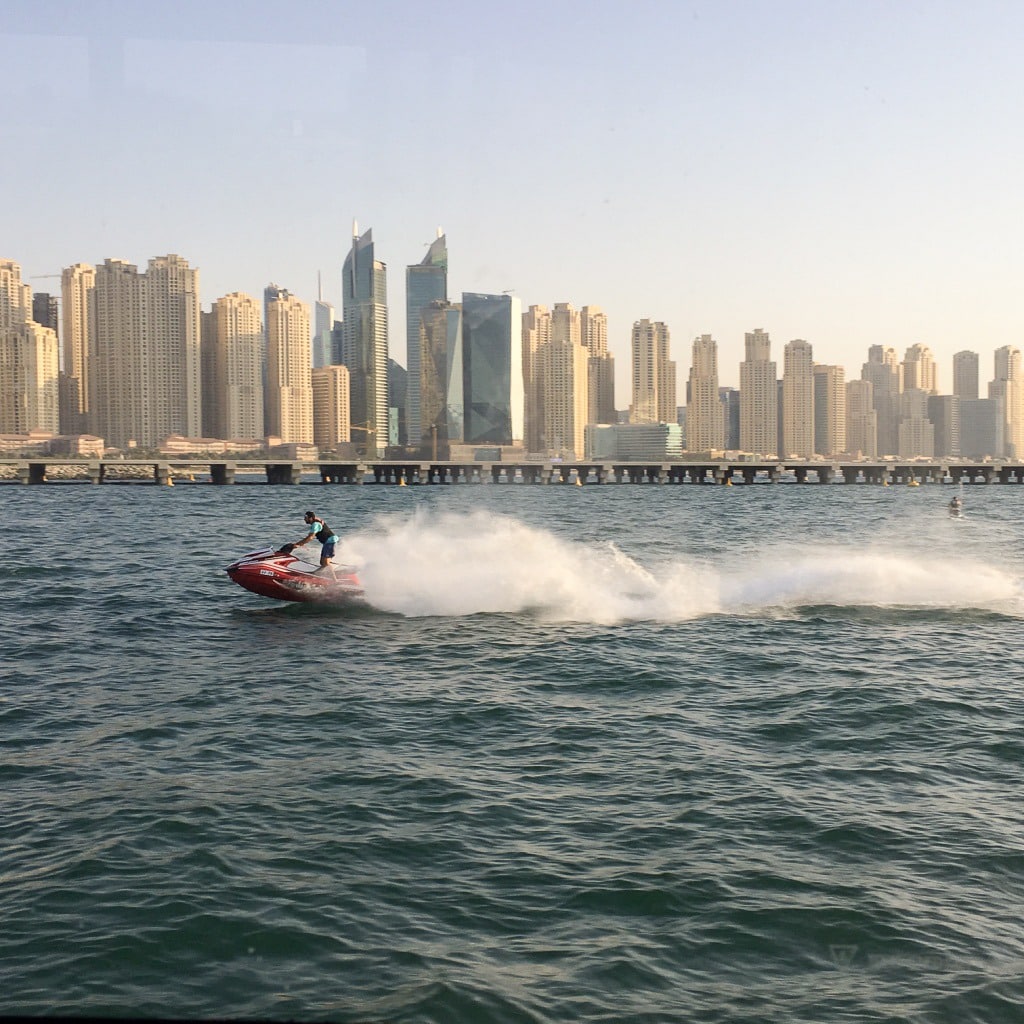 Photo taken across water to the skyscrapers of the Dubai skyline. A man is powering a jet ski at speed across the middle of the picture