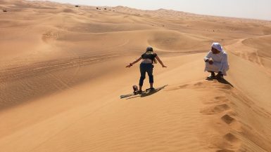Girl sand boarding down steep orange dunes, with man bent down to supervise her