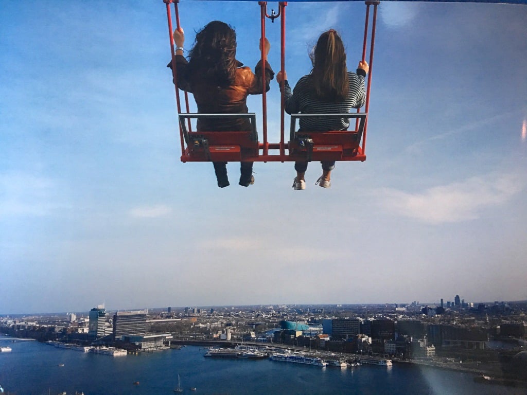 Looking out from the top of a building towards a cityscape below on the other side of a a river. There are two ladies on a red metal swing in the air above the scene. They are both holding on to the sides of the chair and looking away from the camera