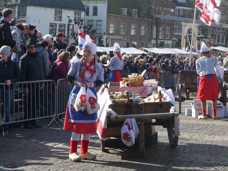 A lady in red, blue and white Dutch outfit wearing clogs, an apron and peaked hat. Stands by a wooden cart with a crowd in the background.