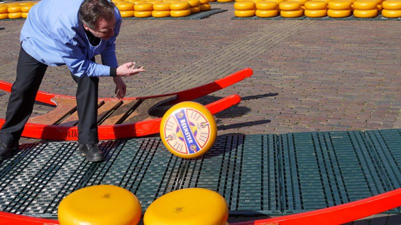 Man wearing pale blue top is rolling a large orange round of cheese along the ground. There are other orange rounds of cheese in the foreground, and stacked in the background