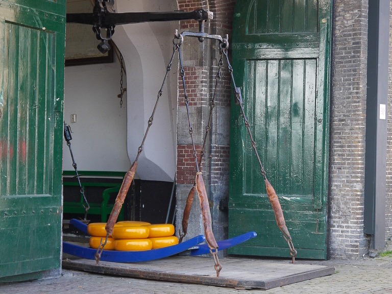 View towards large green wooden doors - open to reveal a large old fashioned scales with metal poles and a wooden base. A blue pallet of round orange cheeses sits on the wooden base.