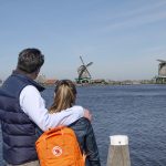 Man in blue shirt and navy gilet with arm around daughter wearing orange rucksack, looking out over the river with windmills in the distance
