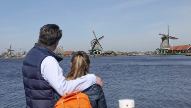 Man in blue shirt and navy gilet with arm around daughter wearing orange rucksack, looking out over the river with windmills in the distance