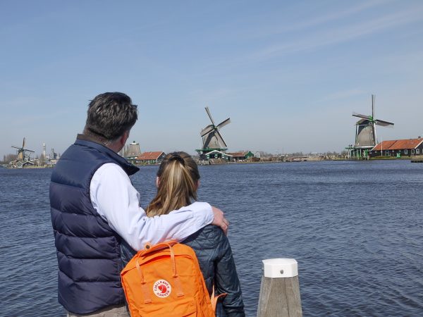 Man in blue shirt and navy gilet with arm around daughter wearing orange rucksack, looking out over the river with windmills in the distance