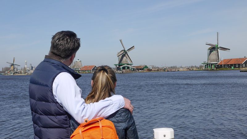 Man in blue shirt and navy gilet with arm around daughter wearing orange rucksack, looking out over the river with windmills in the distance