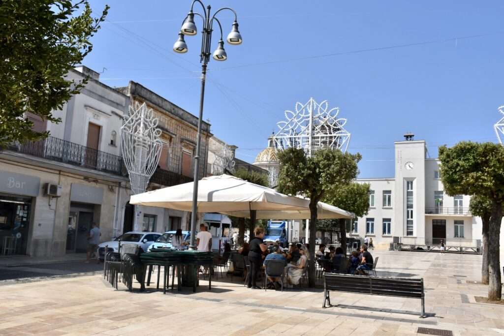 Town square scene with square two storey buildings around the outside and chairs and tables in the middle with diners sitting under a sun shade. Trees to the left, right and middle. Tall silver lamppost with 4 lights to the left. Blue sky above