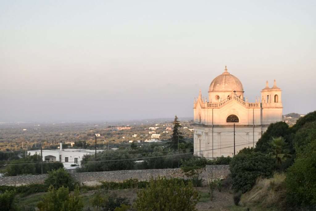 Looking down a hill across a Vally at sun down. A white building with a cupola sits to the right hand side. It is bathed in peachy light. Pale blue sky above