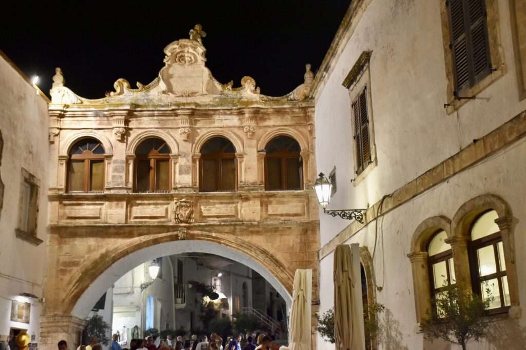 Night time shot of an old, ornately decorated bridge with a large arch at the bottom and row of smaller arches over the top. Stone building to the right hand side