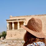 Lady in white dress with long brown hair and a brown straw hat looking away from the camera towards an ancient monument with figures of ladies as columns holding up the roof. Blue sky