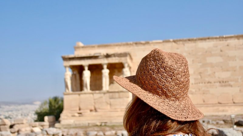 Lady in white dress with long brown hair and a brown straw hat looking away from the camera towards an ancient monument with figures of ladies as columns holding up the roof. Blue sky