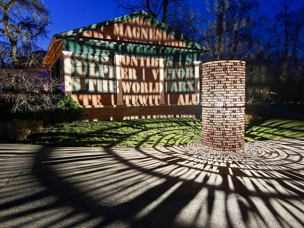 Sculpture lit up at night. Metal cylinder to the right hand side illuminated from the centre and projecting words across the pavement. Building in background behind which has names of water springs projected onto the building