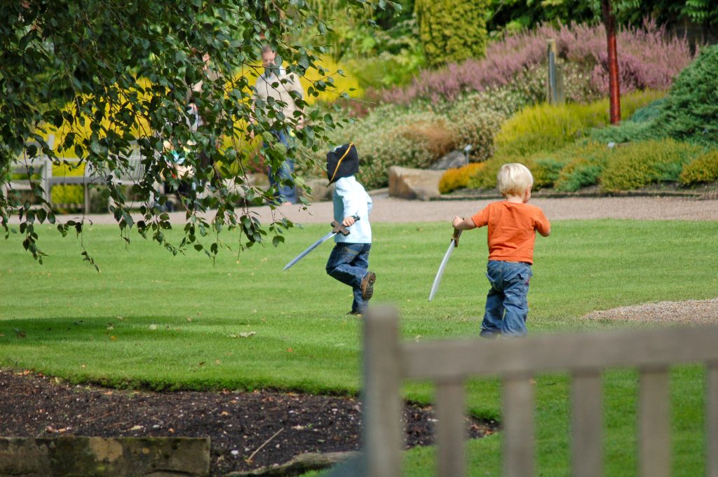 Two young boys playing pirates in the part - carrying plastic swords one in a pirate hat. Photo taken over the back of a bench and with the leaves of a tree overhanging the left of photo
