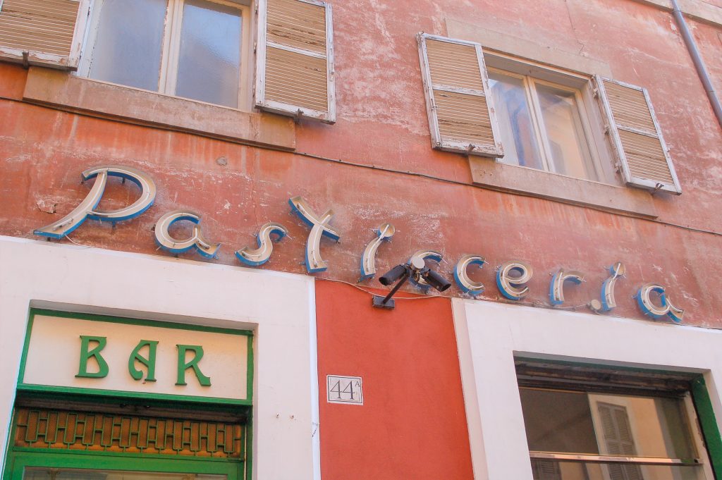 Terracotta coloured wall with white shuttered windows on top floor. Across the centre of photo is a Pasticceria sign with white neon tubing. Pasticceria sign fixed above two windows. The left one has a green BAR sign. There is a No. 44 on the wall