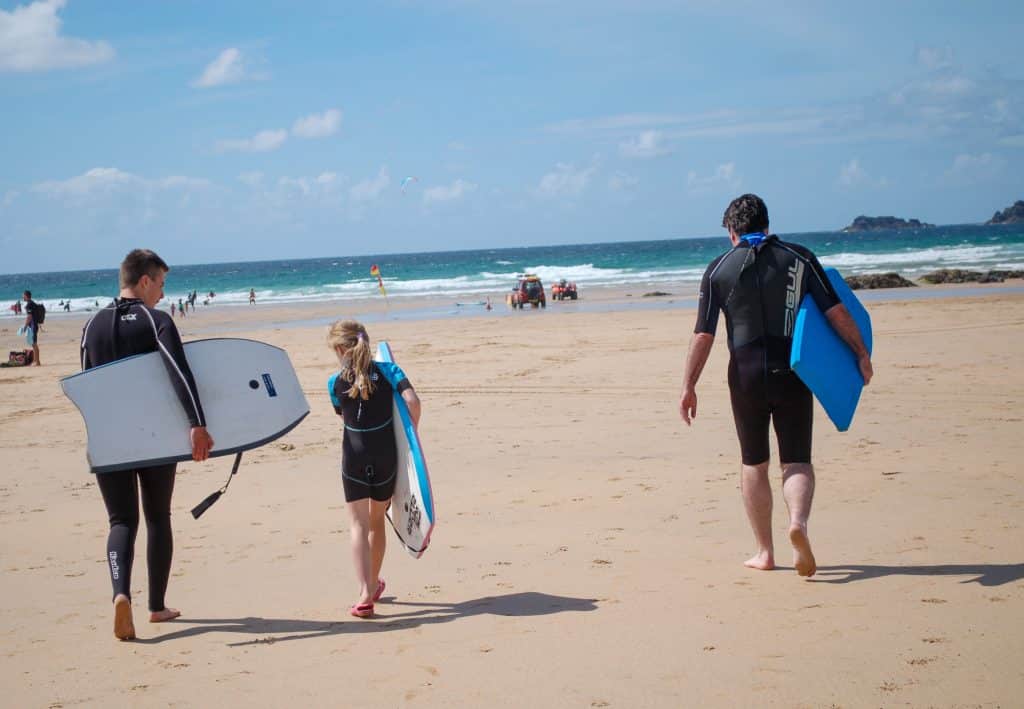 Top third is a pale blue sky with faint grey clouds, bottom two thirds is a wide stretch of sandy beach leading to the sea whose waves you can see on the horizon. A man with teenage boy and girl in wet suits are making their way to the sea with body boards (white and blue)