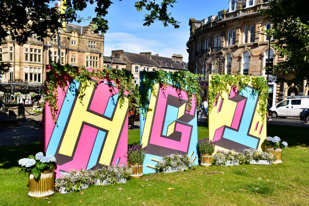 Green grass in foreground and stone buildings in the background. Three multicoloured boards in the middle spelling H, G, 1 (in pink, yellow and blue)