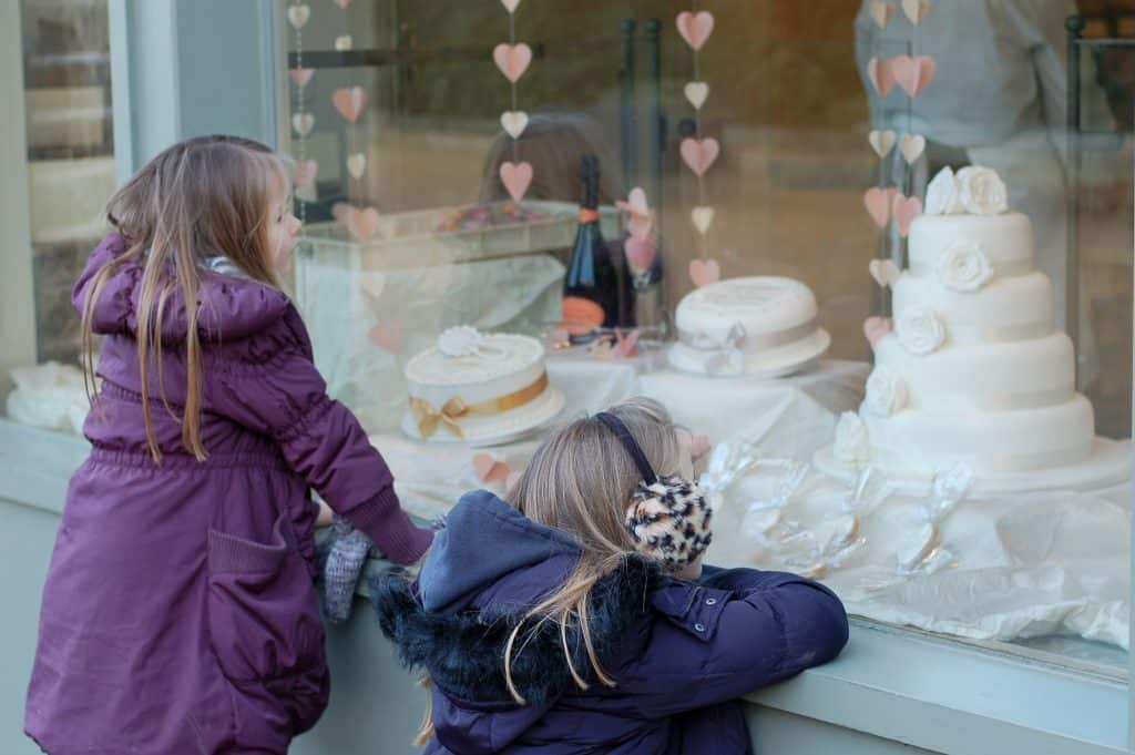 Two young girls looking in the window of a cake shop with white wedding cakes in the window and strings of hearts. The girls are in winter coats - girls on left standing up in purple coat, girl on right kneeling down in navy coat with ear muffs on