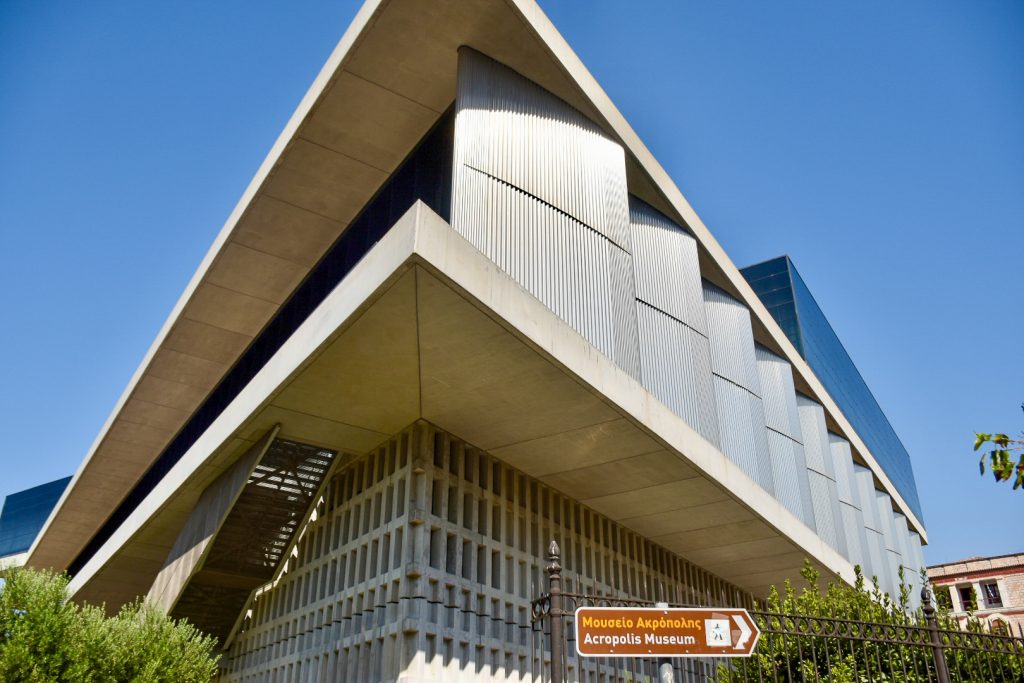 Photo of corner of building facing camera. Modern building with a mix of stone, aluminium and glass on the top. Sign in front pointing to the Acropolis Museum
