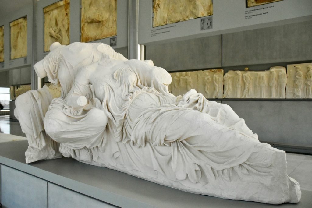 White marble sculputres of two ladies in loose robes reclining away from the camera, one behind the other. Headless and armless. Marble friezes mounted on the walls behind
