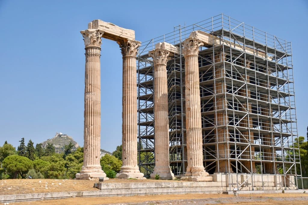 View of 4 large ribbed stone columns with Corinthian detail. The front two are attached to each other by a stone plinth across the top. The two behind are attached to a number of other columns hidden behind scaffolding. Trees to the left hand side and a hilltop in the background