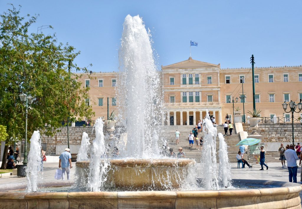 Fountain with one large shoot of water in the middle, surrounded by six smaller shoots of water in a stone basin. Stone palace building with a Greek flag in the background