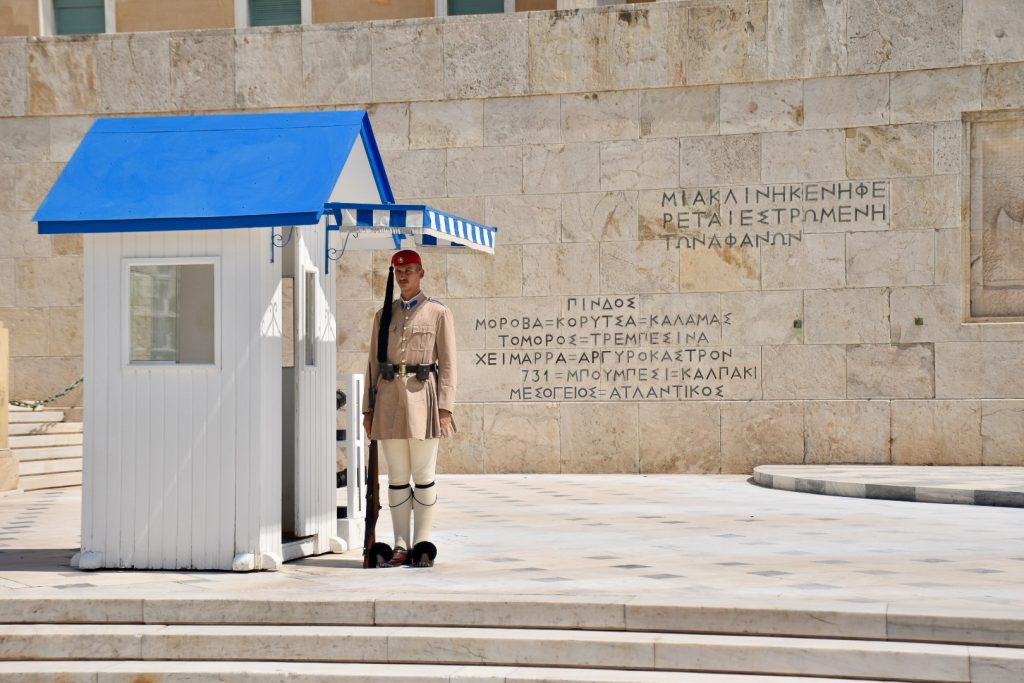 Sentry guard in Greek uniform (black shoes with pompoms, white stockings, beige kilt, beige jacket, belt, berre and gun at side) standing to attention next to white hut with blue roof in front of a stone wall with inscriptions
