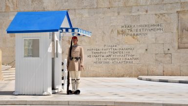 Sentry guard in Greek uniform (black shoes with pompoms, white stockings, beige kilt, beige jacket, belt, berre and gun at side) standing to attention next to white hut with blue roof in front of a stone wall with inscriptions