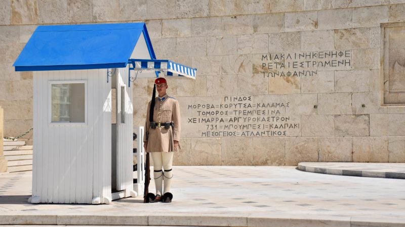 Sentry guard in Greek uniform (black shoes with pompoms, white stockings, beige kilt, beige jacket, belt, berre and gun at side) standing to attention next to white hut with blue roof in front of a stone wall with inscriptions