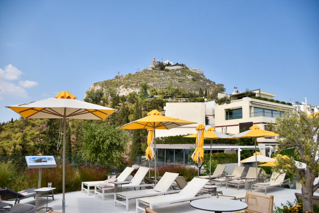 view of pool sun loungers with yellow parasols against a backdrop of a green hill with small church on top
