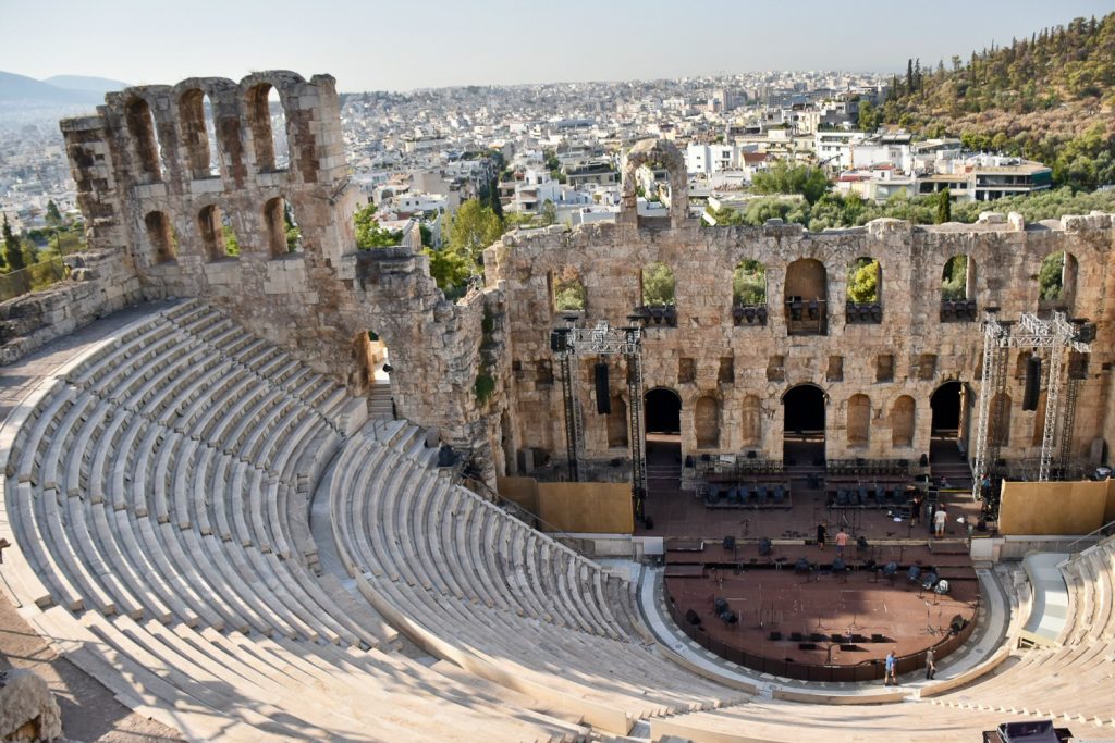 View of a stone amphitheatre spread out on the slope below the photographer. Stone arches at the back of the curved seating area - some intact and some not. In the background, trees to the right and white buildings to to the middle and left