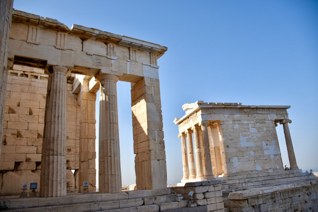 Golden pillars of ancient temples against a pale blue sky. To the left of the photo is part of a square building with two columns. To the right and slightly behind is a square temple with 4 columns at the front and 1 visible on the far side. Flat roof