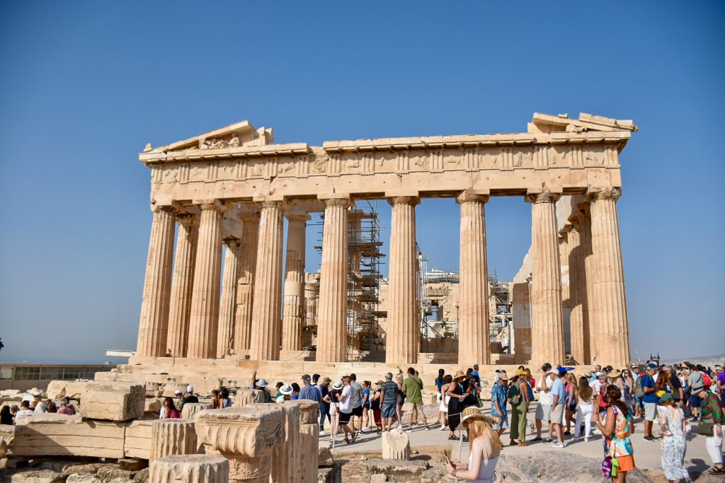 Large sand coloured temple with 8 huge pillars in front and pillars leading backwards along the sides of the building. The remnants of a decorated roof on either sides of the front. Fallen decorative pillars in the foreground. Numerous tourists in front of the ancient temple