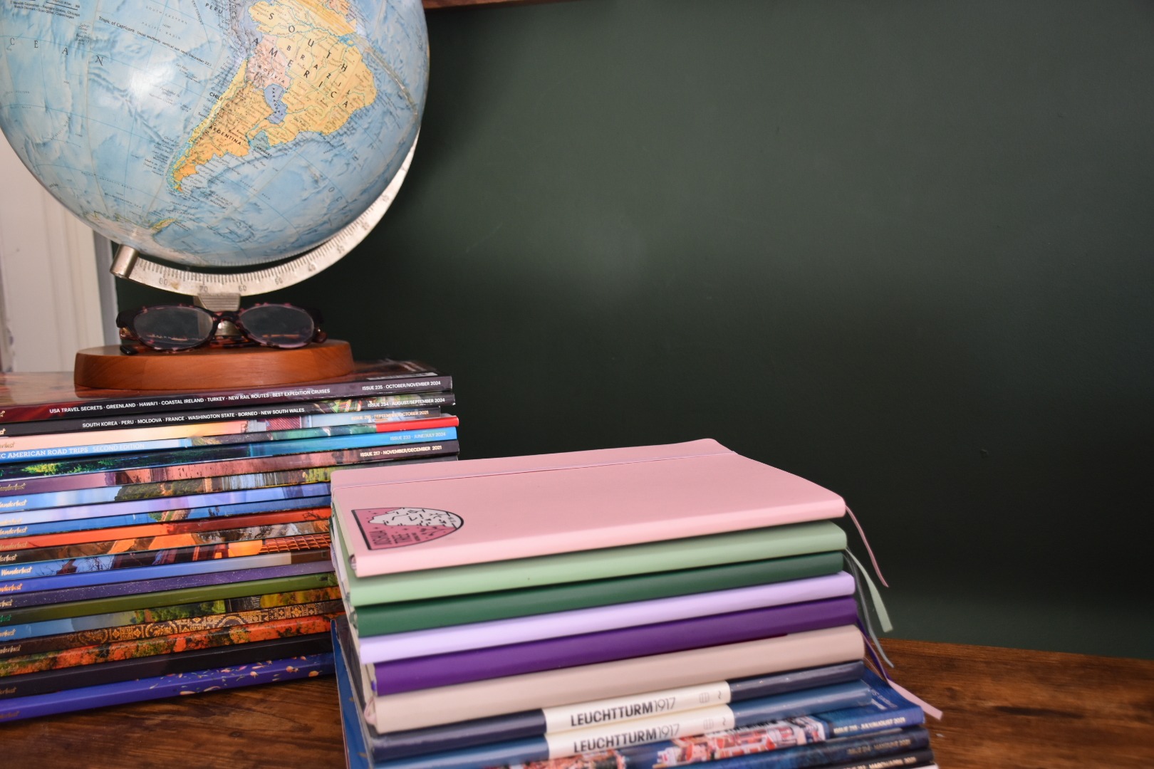 Pile of multicoloured exercise books on a wooden desk with a dark green wall behind. To the far left of the photo a pile of magazines with a globe on top