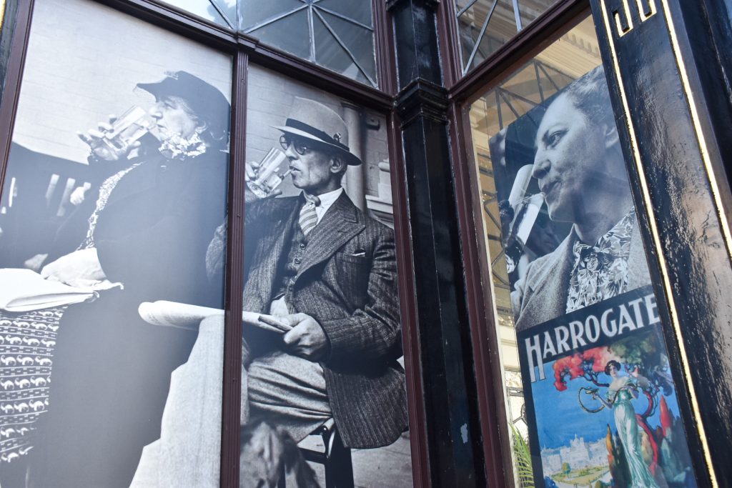 Photo of the side of a building which panels filled in with posters showing 2 ladies and a man in Victorian outfits drinking glasses of water. There is also a posted for Harrogate in the bottom right hand side