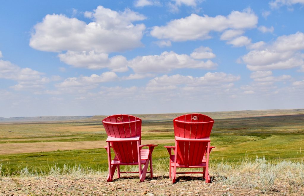Back of two red Adirondack chairs facing out across a wide grassy valley with pale blue sky with white fluffy clouds