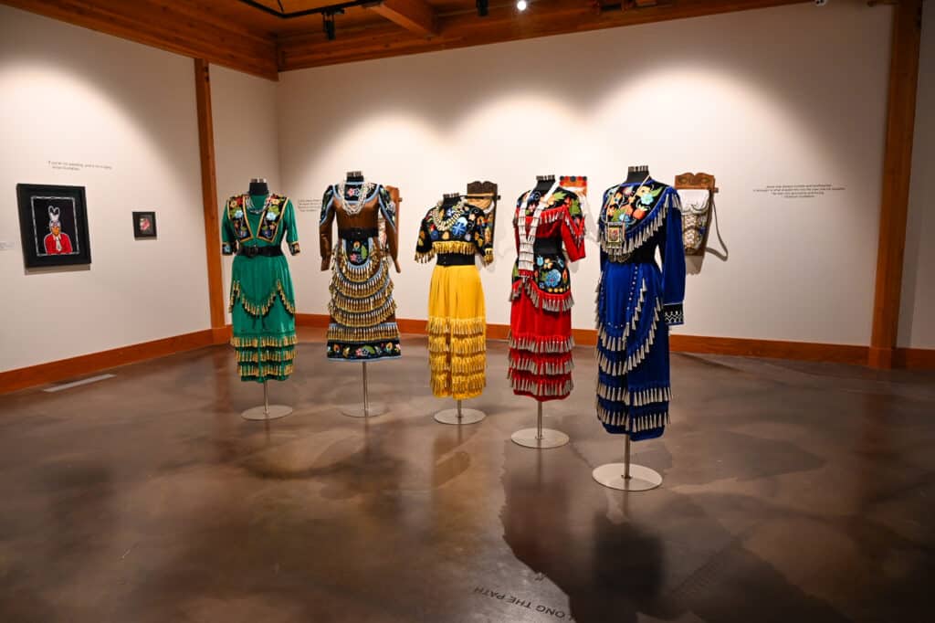 Gallery at Wanuskewin Heritage Park featuring five mannequins dressed in colourful traditional First Nations regalia with floral beadwork, fringe, and layered skirts in green, brown, yellow, red, and blue, displayed under wooden beams on a polished concrete floor