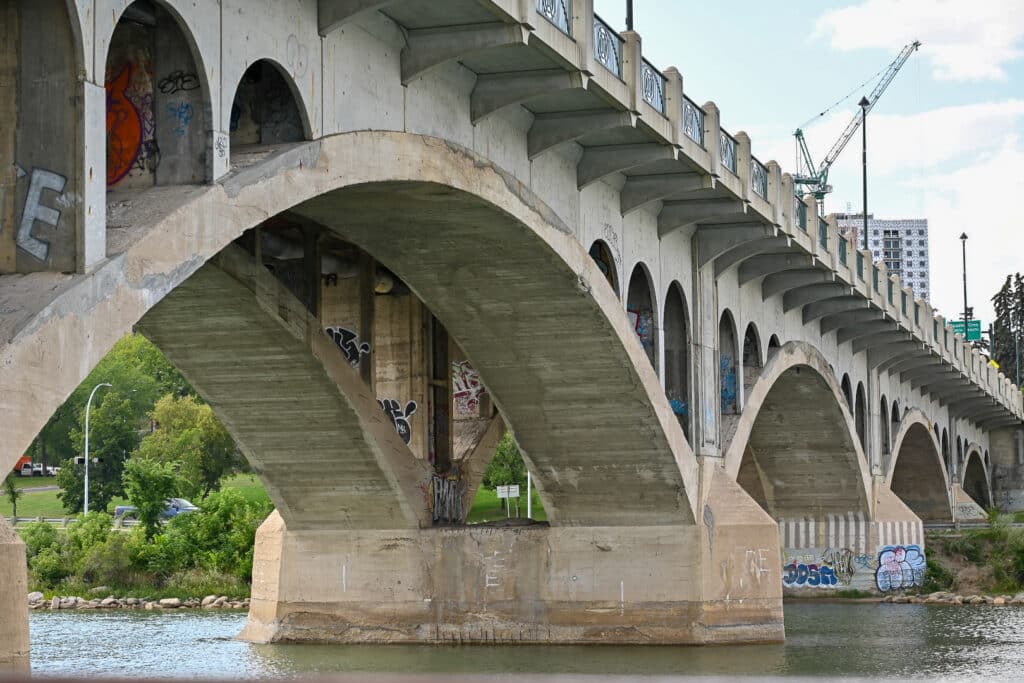 Underside of a multi-arched concrete bridge (Broadway Bridge) spanning the South Saskatchewan River in Saskatoon, showing graffiti-covered supports, ornamental railings, and construction cranes in the background.