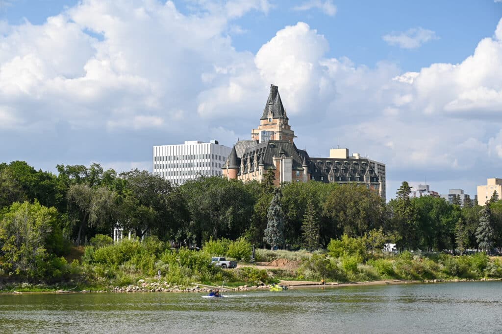 Iconic Château-style Delta Bessborough Hotel rising above dense green trees along the South Saskatchewan River in Saskatoon, with people and a jet ski on the water under a partly cloudy blue sky.
