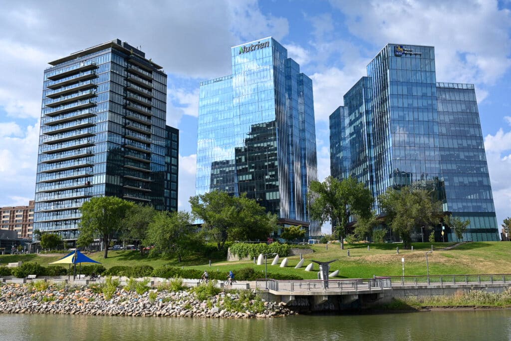 Modern glass office towers including the Nutrien and RBC Wealth Management buildings along the South Saskatchewan River in downtown Saskatoon, with a landscaped riverbank, pathway, and cyclists in the foreground.