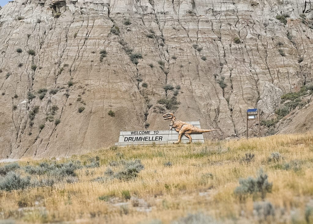Large “Welcome to Drumheller” sign featuring a life-sized golden Tyrannosaurus rex statue standing against layered beige badlands cliffs, with dry yellow grass and sagebrush in the foreground under a clear sky.
