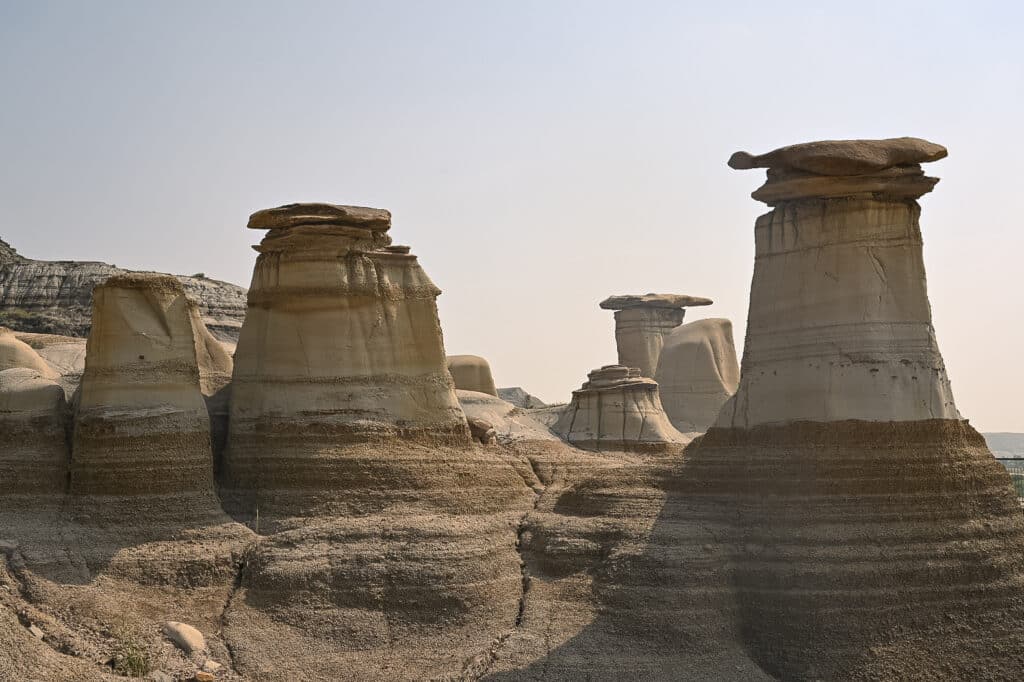 Dramatic hoodoo rock formations in the Alberta Badlands near Drumheller — tall, eroded sandstone pillars with flat capstones, layered in beige and gray tones under a hazy pale sky.