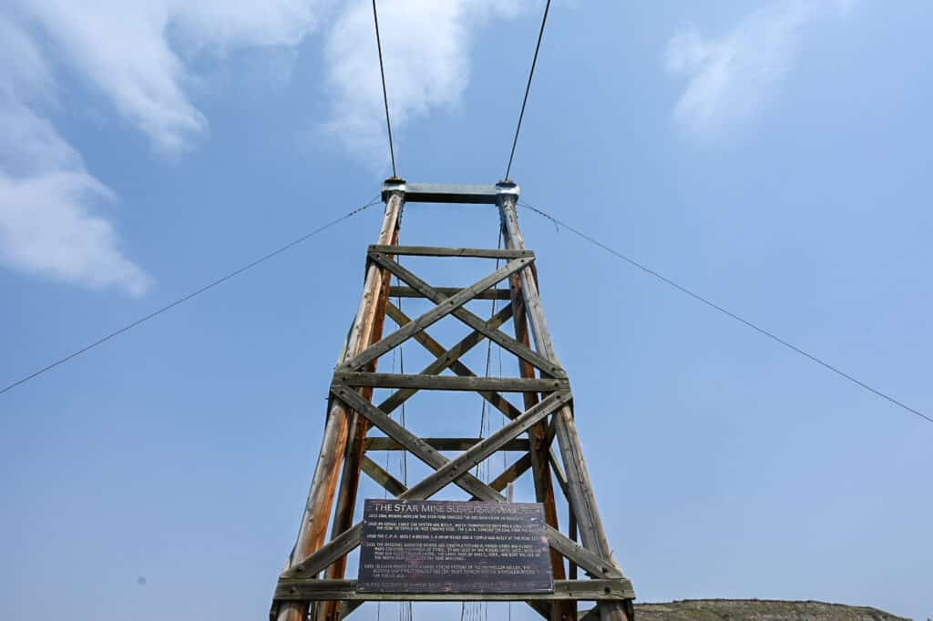 Looking up at the wooden A-frame tower of the Star Mine Suspension Bridge near Drumheller against a bright blue sky with light clouds; a historical plaque is mounted at the base of the structure.
