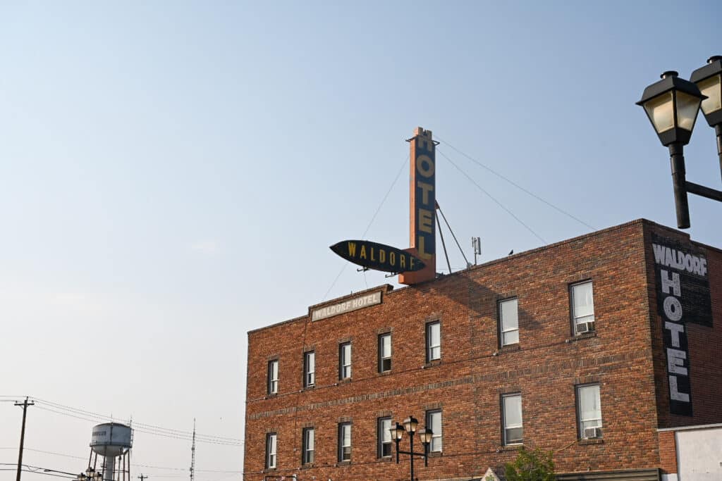 Red-brick Waldorf Hotel in downtown Drumheller, Alberta, featuring a tall vertical “HOTEL” sign, a classic black-and-yellow “WALDORF” arrow sign, and a painted “WALDORF HOTEL” mural on the side wall; a Drumheller water tower is visible in the background against a clear blue sky.