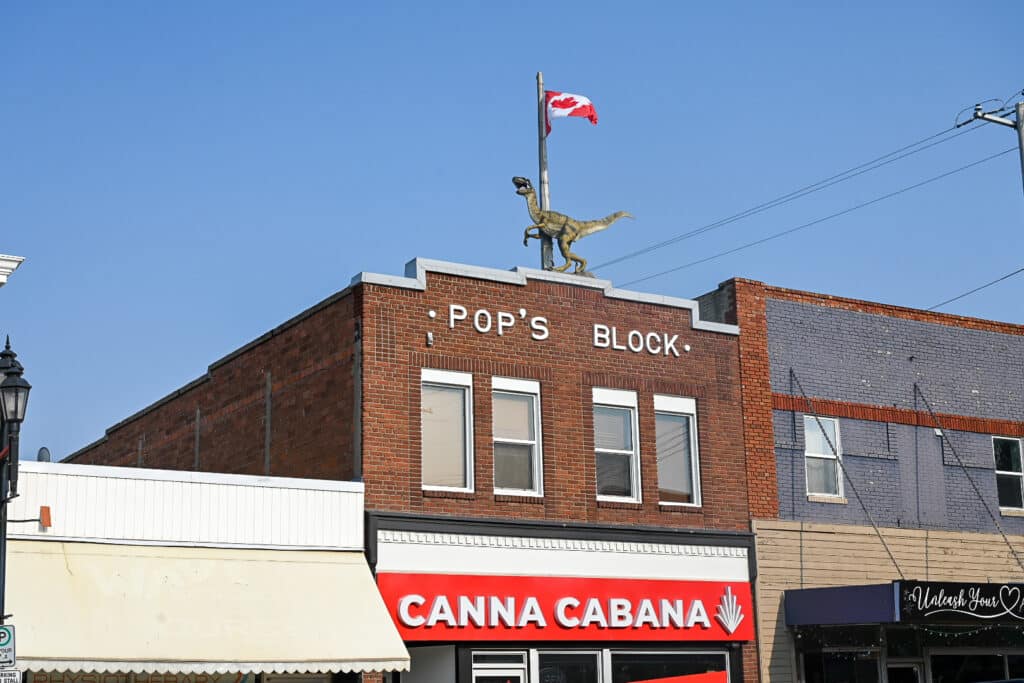 Brick commercial building labeled “Pop’s Block” in downtown Drumheller, topped with a life-sized dinosaur statue and a Canadian flag flying against a clear blue sky; storefronts including Canna Cabana are visible below.