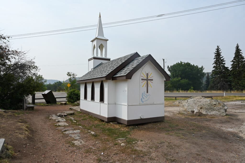 Small white wooden roadside chapel with a steeple, brown trim, arched windows, and a painted cross with radiating rays and a dove on the side wall, situated beside a highway amid trees, rocks, and dry grass near Drumheller.
