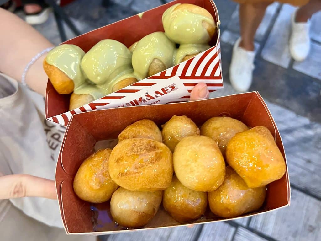 Hand holding two open cardboard boxes of doughnut balls towards the camera. The bottom box has a red inside and is filled with doughtnut balls glistening in honey. The bank box with red and white stripey design is filled with doughnut balls covered in green runny sauce (pistachio)