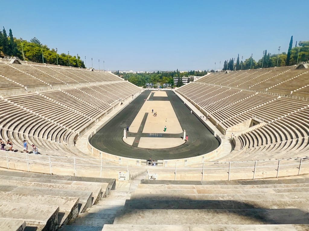 View of a white marble running track taken from the far end looking down the dark track from the upper tiers of the stadium. Lights on the top of the stadium and trees seen from beyond the stadium walls