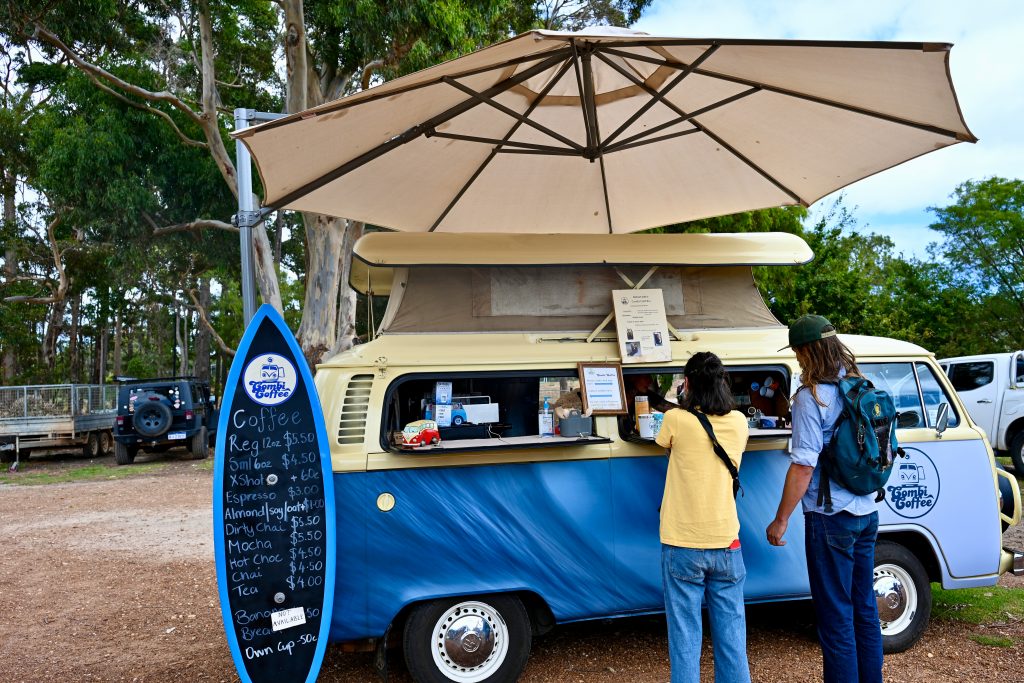 A blue VW Camper van with a cream roof sits in the middle of the frame. It is a coffee bar and a surfboard shaped notice board advertises the menu to the left. A large beige parasol sits above the van. A lady in yellow t-shirt and jeans, and a man in denim with long hair and a green baseball hat are ordering at the van window. Trees behind Margaret River shops
