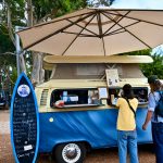 A blue VW Camper van with a cream roof sits in the middle of the frame. It is a coffee bar and a surfboard shaped notice board advertises the menu to the left. A large beige parasol sits above the van. A lady in yellow t-shirt and jeans, and a man in denim with long hair and a green baseball hat are ordering at the van window. Trees behind Margaret River shops